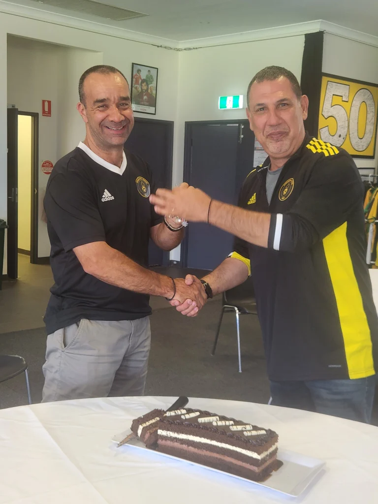 Two committee members shaking hands beside a celebration cake at a club function.