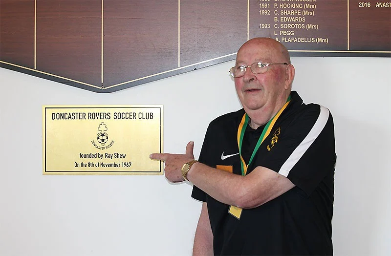 Club founder Ray Shew, wearing club colours and a medal, pointing to a plaque reading “Doncaster Rovers Soccer Club, founded by Ray Shew on the 8th of November 1967”. Club honour boards hang on the wall behind him.